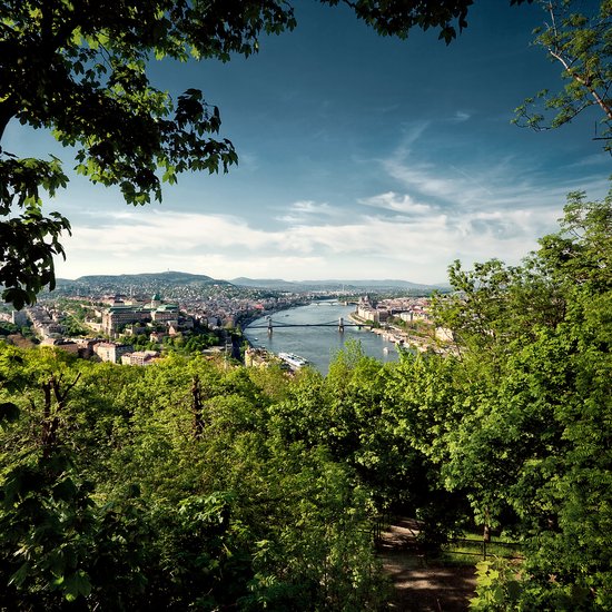 Panoramic view of Budapest from a green park overlooking the bridge over the Danube and the city with a hilly landscape in the background.