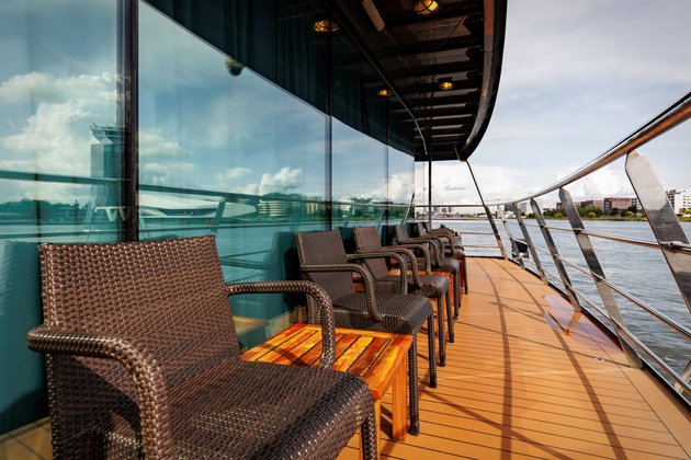 Several armchairs and wooden tables are located in the outdoor area at the stern of the ship, overlooking the river and the city in the background.