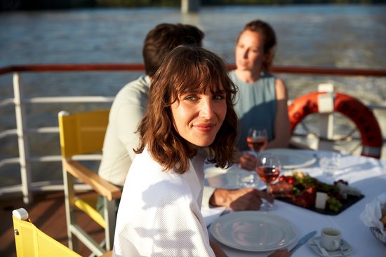 A woman with brown hair and a white sweater smiles on a boat, with two other people and a set table with wine and snacks in the background.