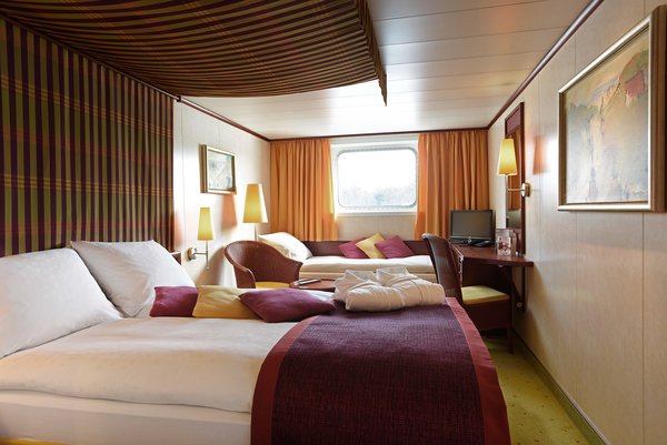 A category A cabin on a Danube ship with a double bed and an additional bed, as well as a window, seating options, and a desk with a television.