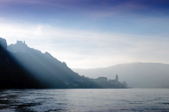 A mystical river landscape in the Wachau with hills on the riverbank. At the foot of the hill lies a village and at the top stands a castle. The sun shines over the hills onto the river.
