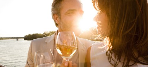 A couple stands at sunset with a glass of wine in hand at the railing of a riverboat. In the background, the river and a riverside landscape.
