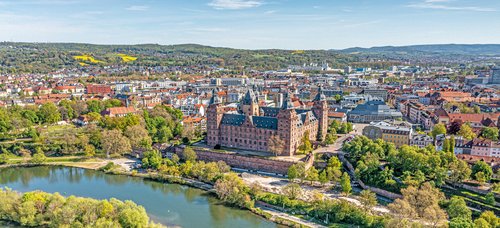 Aschaffenburgs Schloss Johannisburg spiegelt sich im Main, umgeben von grünen Bäumen und einem klaren blauen Himmel.