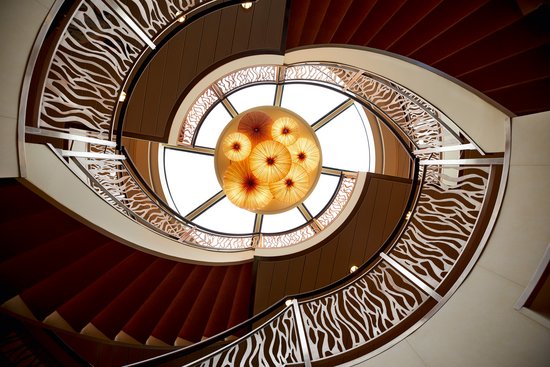 Spiral staircase with decorative railing and round lampshades in the atrium of a ship.
