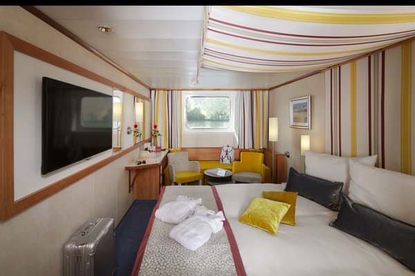 A category A cabin on the A-ROSA DONNA with a double bed, an additional bed, and a window, as well as seating options, a desk, and a flat-screen TV, with luggage and bathrobes in the cabin.