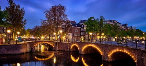 Illuminated stone bridge over a canal in Amsterdam at dusk with historic houses and trees in the background