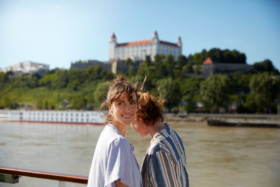 Two women stand at the railing on a river cruise ship on the Danube, looking at another ship and Bratislava Castle on a wooded hill in the background.