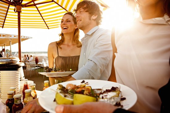 Three people are standing at the buffet at the barbecue on the sun deck of a river cruise ship with grilled food, with the river and trees in the background.